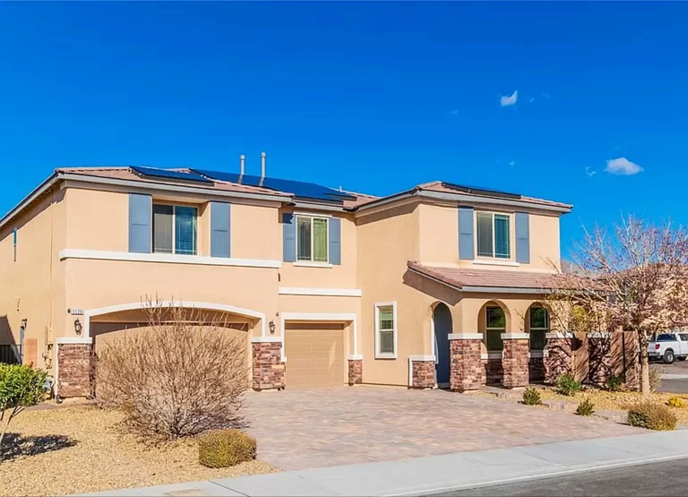 Two-story tan and brick home with solar panels and paved driveway under clear blue sky.