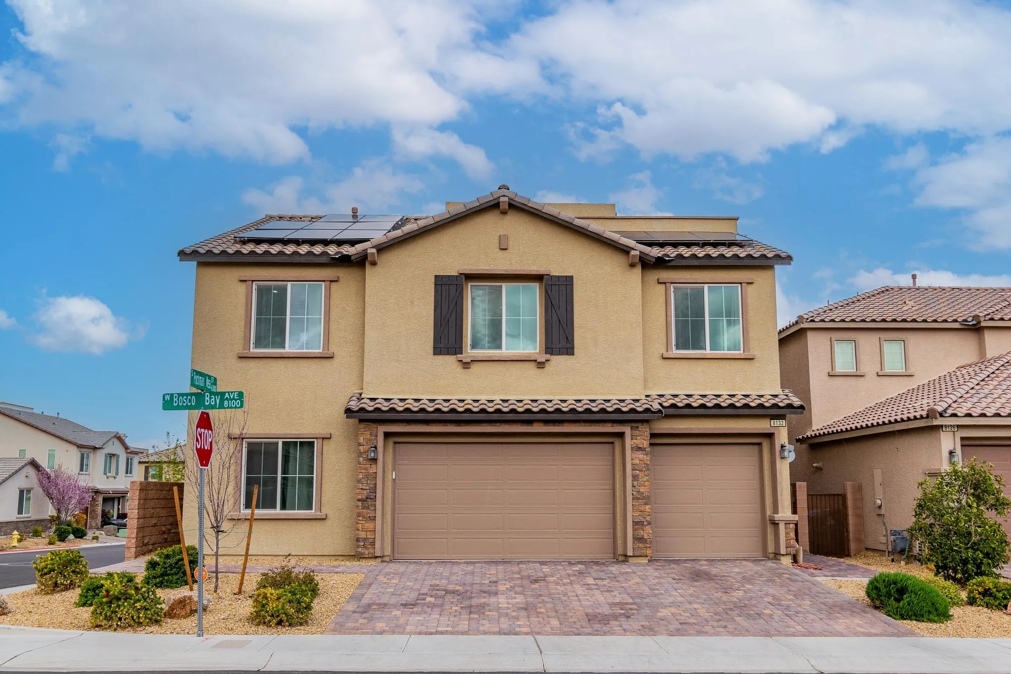 Two-story tan stucco house with brick garage and solar panels on roof in residential neighborhood.